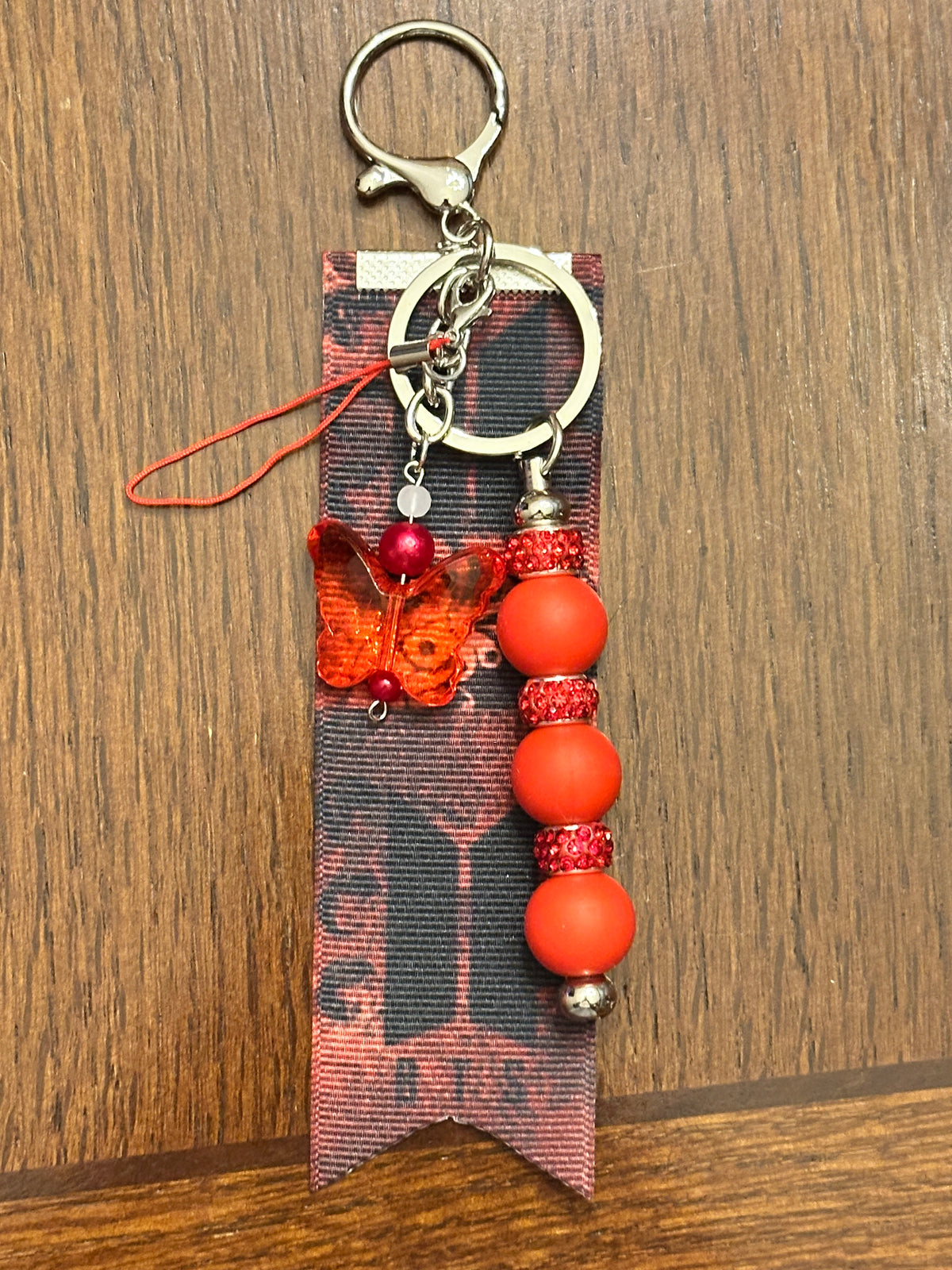 A handmade keychain featuring a silver split ring, a butterfly charm with red beads, a black and red patterned ribbon, and a dangling strand of silicone beads with jeweled spacers