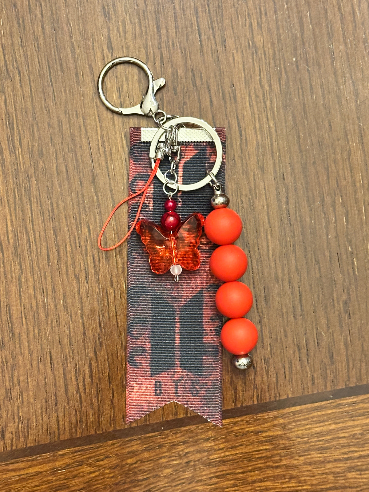 A handmade keychain featuring a silver split ring, a butterfly charm with red beads, a black and red patterned ribbon, and a dangling strand of silicone beads.