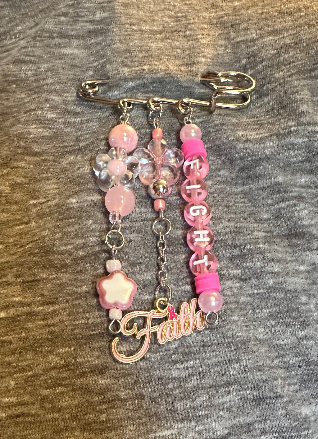 A pink and silver safety pin accessory with dangling charms, including assorted pink beads, clear beads, a flower beads, a letter charm spelling 'FIGHT', a pink 'Faith' script charm, and a pink cylindrical bead accent.
